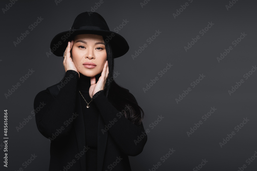 Stylish asian woman in fedora hat touching face isolated on dark grey Stylish asian woman in fedora hat touching face isolated on dark grey