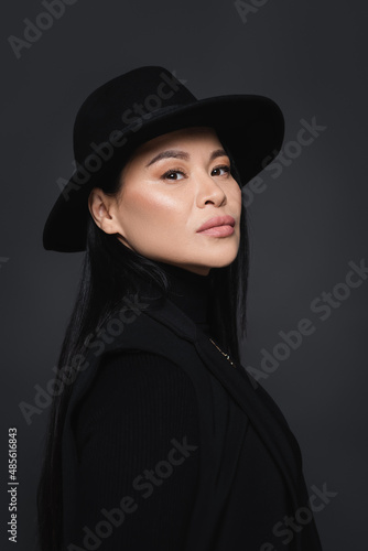 Portrait of asian model in jacket and fedora hat looking at camera isolated on dark grey © LIGHTFIELD STUDIOS