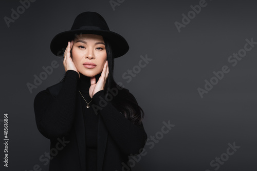 Stylish asian woman in fedora hat touching face isolated on dark grey
