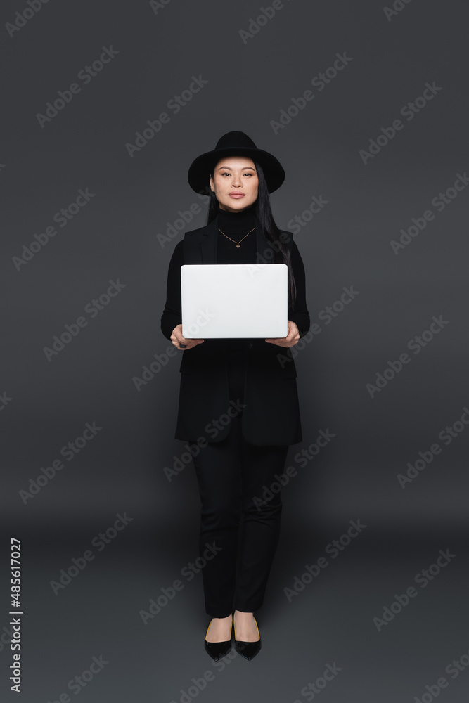 Full length of stylish asian woman in fedora hat holding laptop on dark grey background Full length of stylish asian woman in fedora hat holding laptop on dark grey background