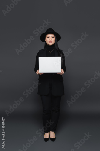 Full length of stylish asian woman in fedora hat holding laptop on dark grey background