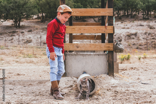 Caucasian village boy in boots and a beret playing with a kitten on a farm