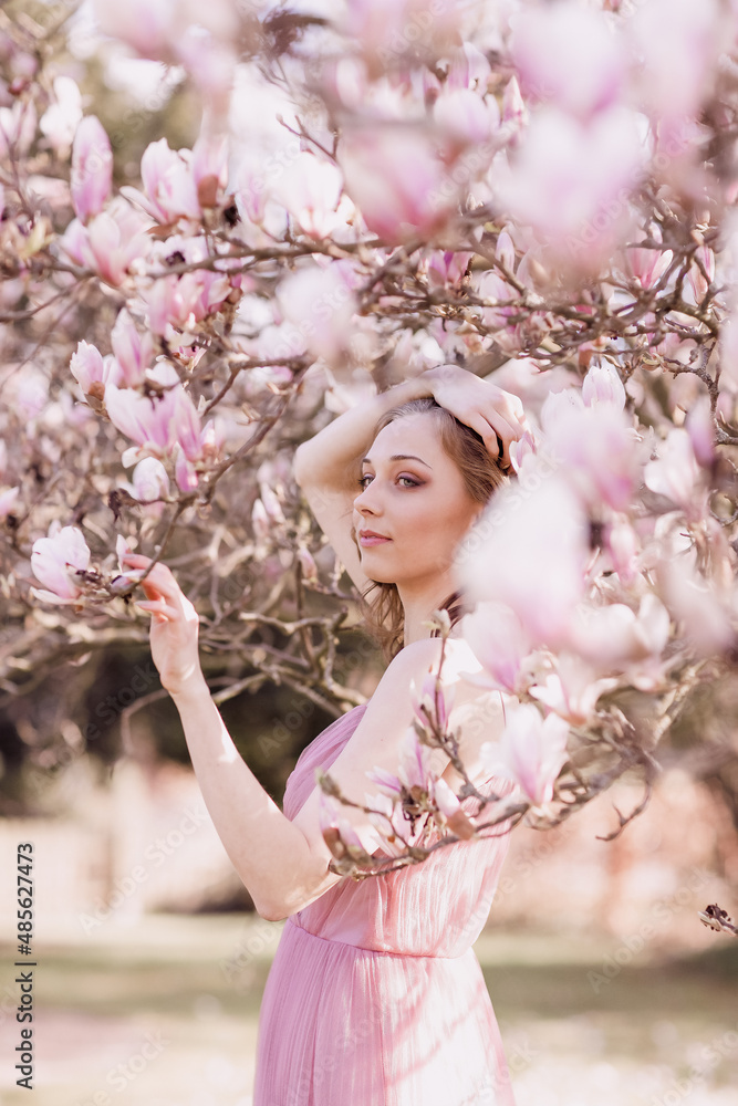 Spring flowering. Portrait of a young blonde girl in the branches of a blooming magnolia. Open aperture with light blurring and illumination. Spring flowering. Portrait of a young blonde girl in the branches of a blooming magnolia. Open aperture with light blurring and illumination.