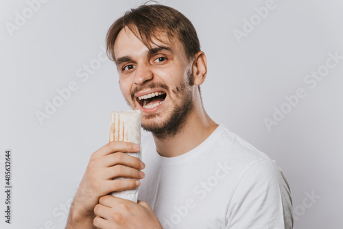 A young man in a clean T-shirt on a white background holds lunch in his hands, shawarma with chicken.