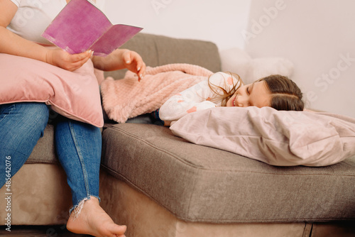 Cropped photo of a mother sitting on the sofa and holding a book reading a fairy tale for her sleeping daughter in the evening,