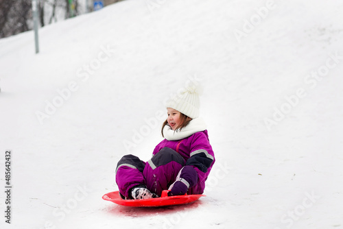 Cute funny child girl in a purple overalls and a hat with a pompon in the park rides on a bright red ice rink during a snowfall. Winter outdoor activities