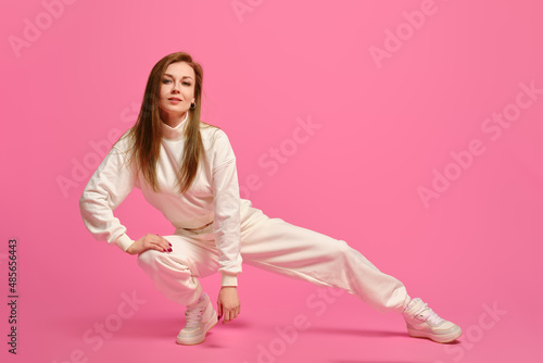 Active woman in white sportswear sits on squats and leaning on one leg over pink background