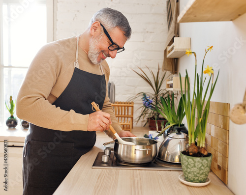 Photography Happy senior man in apron cooking in kitchen at home, standing near stove and st