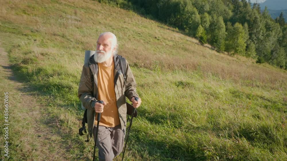 Happy Elderly Man with a Backpack and Nordic Walking Poles Walks Along the Mountain Trail. The Traveler Makes a Hike in the Mountains, Enjoys Nature. Adventure Tourism, Outdoor Activities and Hobbies. Happy Elderly Man with a Backpack and Nordic Walking Poles Walks Along the Mountain Trail. The Traveler Makes a Hike in the Mountains, Enjoys Nature. Adventure Tourism, Outdoor Activities and Hobbies.