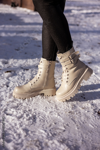 Woman in fashionable white boots on white snow, close-up. Women's legs in stylish winter leather boots