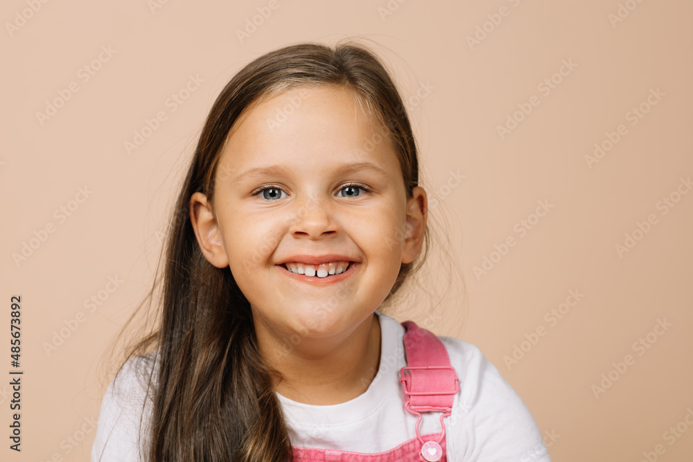 Portrait of child with bright shining eyes and excited happy smile with upper teeth looking at camera wearing bright, pink jumpsuit and white t-shirt on beige background. Portrait of child with bright shining eyes and excited happy smile with upper teeth looking at camera wearing bright, pink jumpsuit and white t-shirt on beige background.