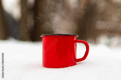 Red enamel mug cup coffee hot drink in snow against winter forest background. Close up. Copy space