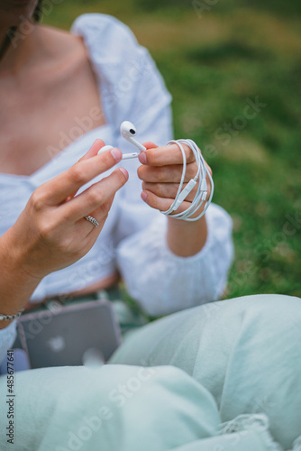 A teenager wrapping the ear buds wire after listening to music. She is sitting on the grass in a park.
