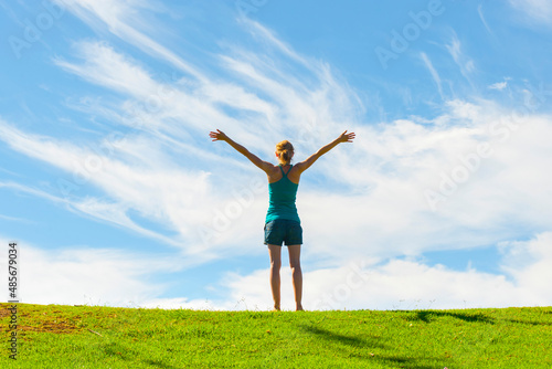 Happy woman raising arms up in air with joy, enjoying happiness of freedom and carefree lifestyle on green grass with blue sky, background with copy space