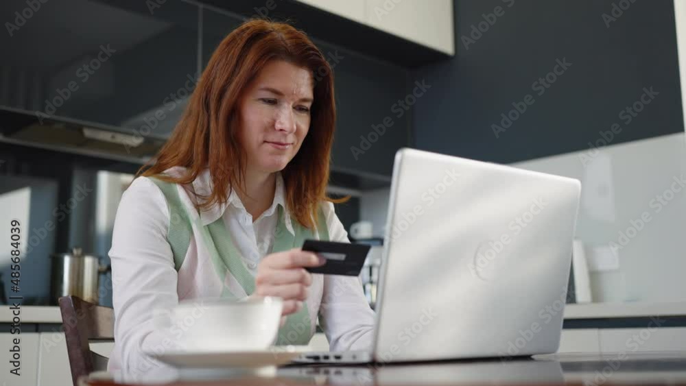 Adult woman making online order with credit card while sitting in kitchen, using application on laptop, showing thumb up, making yes gesture. Concept of shopping Adult woman making online order with credit card while sitting in kitchen, using application on laptop, showing thumb up, making yes gesture. Concept of shopping