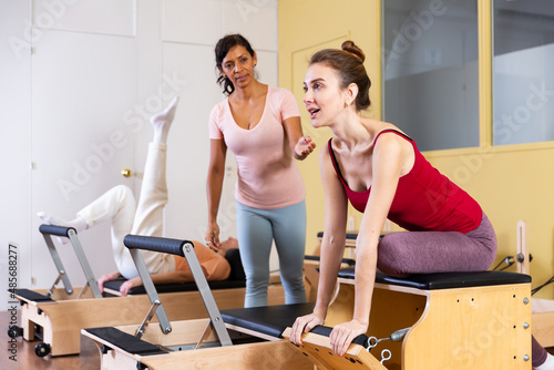 Young active woman practicing Pilates in a group workout with an instructor performs an exercise on a combined chair in a ..fitness studio
