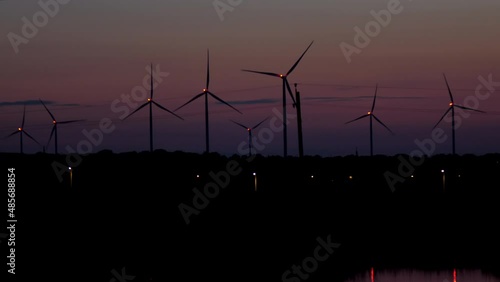 Wind turbines generate renewable energy at night. Silhouettes of windmills with bright lights on propellers rotate against dark sky near river photo