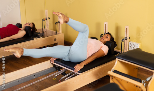 Active latin american woman practicing Pilates in a fitness studio performs a stretching exercise on a reformer bed