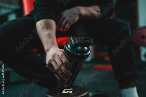 Close up on hand of unknown caucasian man holding dark supplement shaker while sitting at gym during training copy space selective focus photo
