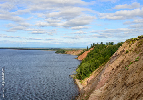 View of Andoma cliff on the eastern coast of the Onega lake, Vologda region, northern Russia. Deep blue water, light blue sky with clouds, green trees, highh brown bank