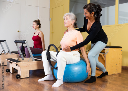 Senior woman doing exercises with resistance ring and fitball ​in pilates studio under supervision of qualified Hispanic female trainer..