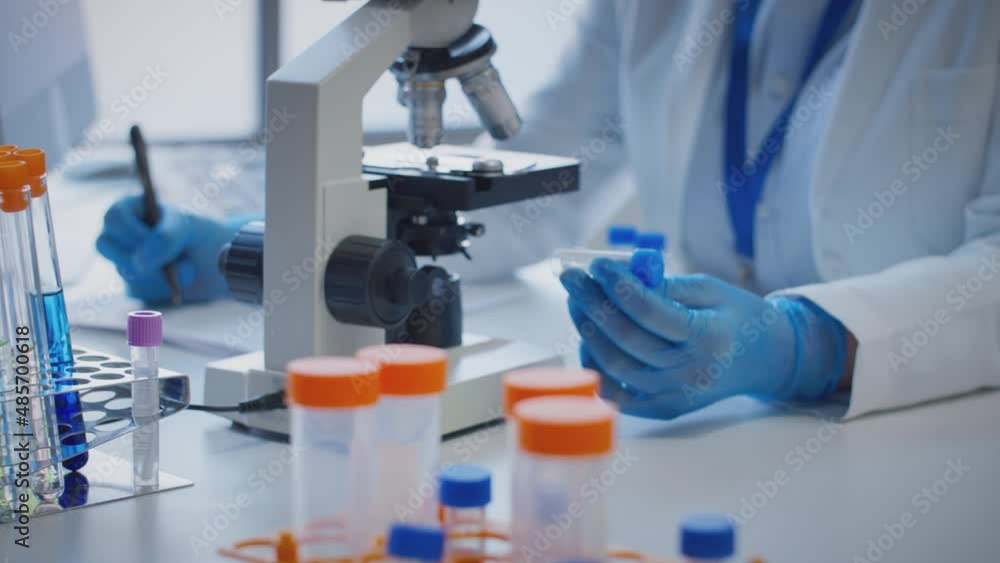 Close up of scientist wearing white coat looking through microscope and making notes whilst holding test tube which he puts on desk to show label lambda - shot in slow motion Close up of scientist wearing white coat looking through microscope and making notes whilst holding test tube which he puts on desk to show label lambda - shot in slow motion