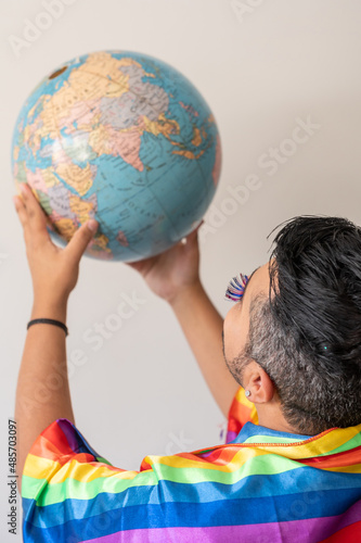 LGBT man boy with ranbow flag holding earth globe, wearing lashes, smiling and posing forthe camera by a grey background in a studio.  photo