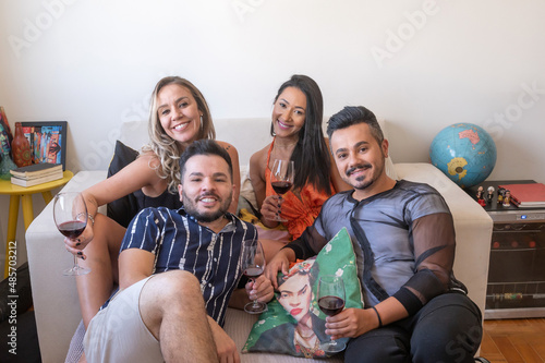 Four LGBT people drinking wine, posing and smiling at the camera, two women and two men, on the couch in a living room, during the day. photo