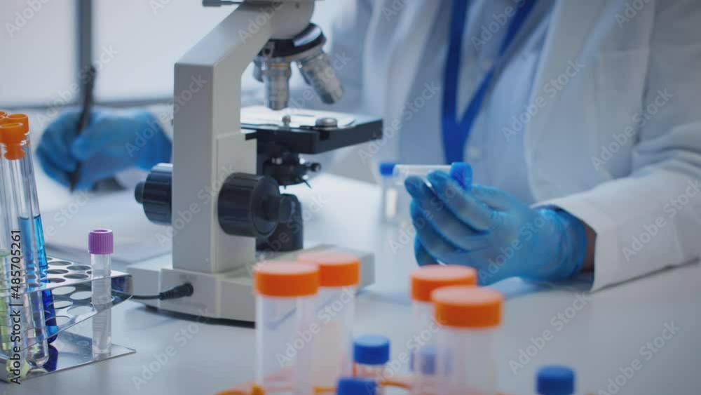 Close up of scientist wearing white coat looking through microscope and making notes whilst holding test tube which he puts on desk to show label lota - shot in slow motion Close up of scientist wearing white coat looking through microscope and making notes whilst holding test tube which he puts on desk to show label lota - shot in slow motion