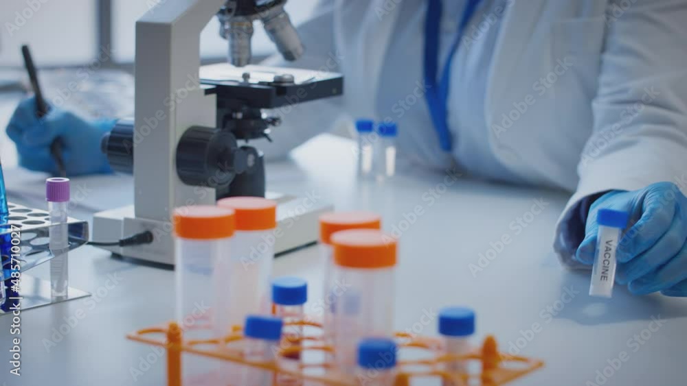 Close up of scientist wearing white coat looking through microscope and making notes whilst holding test tube which he puts on desk to show label vaccine - shot in slow motion Close up of scientist wearing white coat looking through microscope and making notes whilst holding test tube which he puts on desk to show label vaccine - shot in slow motion