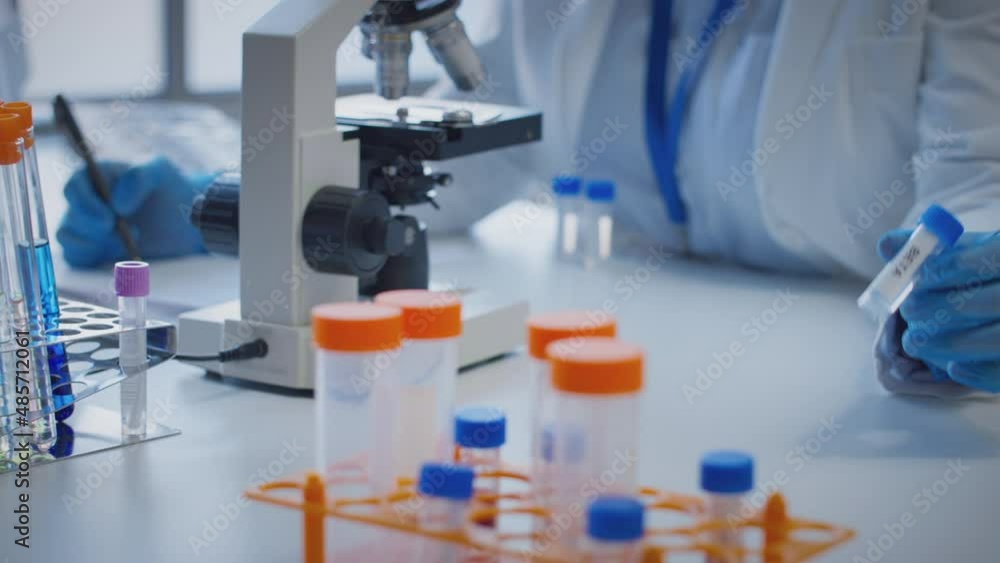 Close up of scientist wearing white coat looking through microscope and making notes whilst holding test tube which he puts on desk to show label beta - shot in slow motion Close up of scientist wearing white coat looking through microscope and making notes whilst holding test tube which he puts on desk to show label beta - shot in slow motion