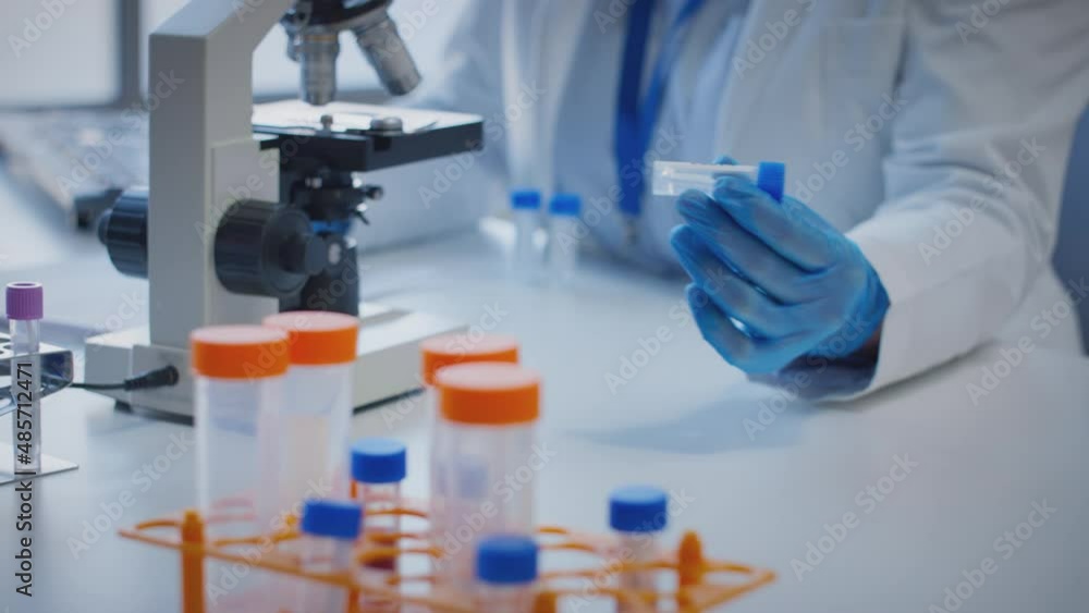Close up of scientist wearing white coat looking through microscope and making notes whilst holding test tube which he puts on desk to show label kappa - shot in slow motion Close up of scientist wearing white coat looking through microscope and making notes whilst holding test tube which he puts on desk to show label kappa - shot in slow motion