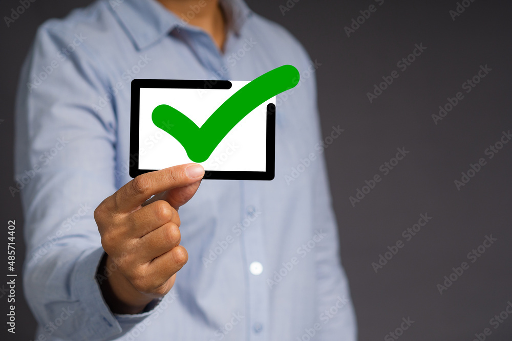 Hand holding a green check mark symbol with a black border while standing against a gray background. Hand holding a green check mark symbol with a black border while standing against a gray background.