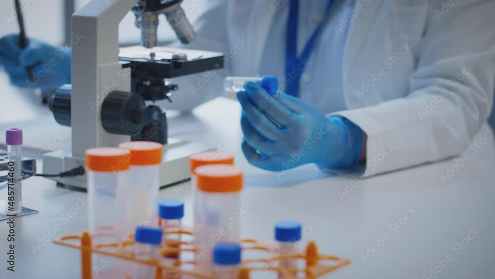 Close up of scientist wearing white coat looking through microscope and making notes whilst holding test tube which he puts on desk to show label Covid-19 - shot in slow motion Close up of scientist wearing white coat looking through microscope and making notes whilst holding test tube which he puts on desk to show label Covid-19 - shot in slow motion