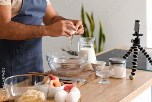 Young man breaking egg into bowl while following cooking video tutorial in kitchen, closeup