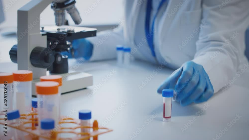 Close up of scientist wearing white coat looking through microscope and making notes whilst holding blood sample in test tube which he puts on desk to show blank label - shot in slow motion Close up of scientist wearing white coat looking through microscope and making notes whilst holding blood sample in test tube which he puts on desk to show blank label - shot in slow motion