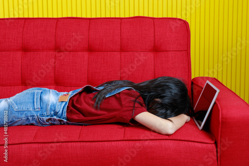 Studio shot Asian young primary school girl in denim jeans overalls outfit laying down sweet dream sleeping on red cozy sofa holding hugging touchscreen tablet computer in arms in yellow living room