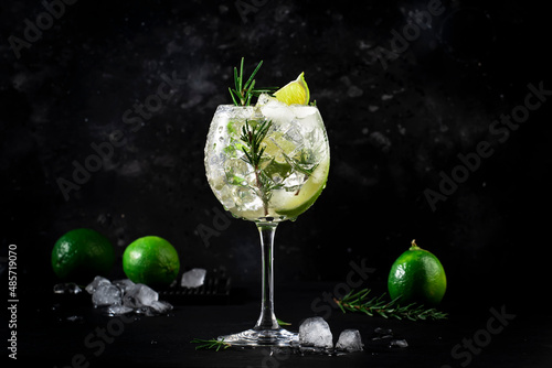 Gin tonic alcoholic cocktail drink with dry gin, rosemary, tonic, lime and ice cubes in wine glass. Black bar counter background, bar tools, copy space photo