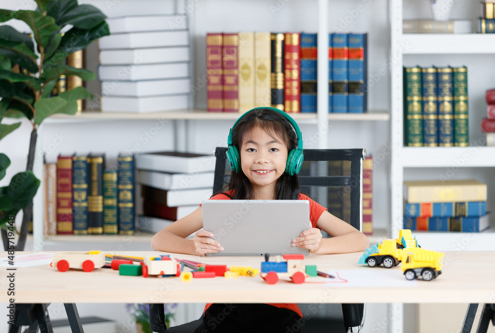 Asian clever smart diligent young primary school girl daughter in casual outfit wearing big headphone smiling using touchscreen tablet computer in hands learning studying online during quarantine Asian clever smart diligent young primary school girl daughter in casual outfit wearing big headphone smiling using touchscreen tablet computer in hands learning studying online during quarantine