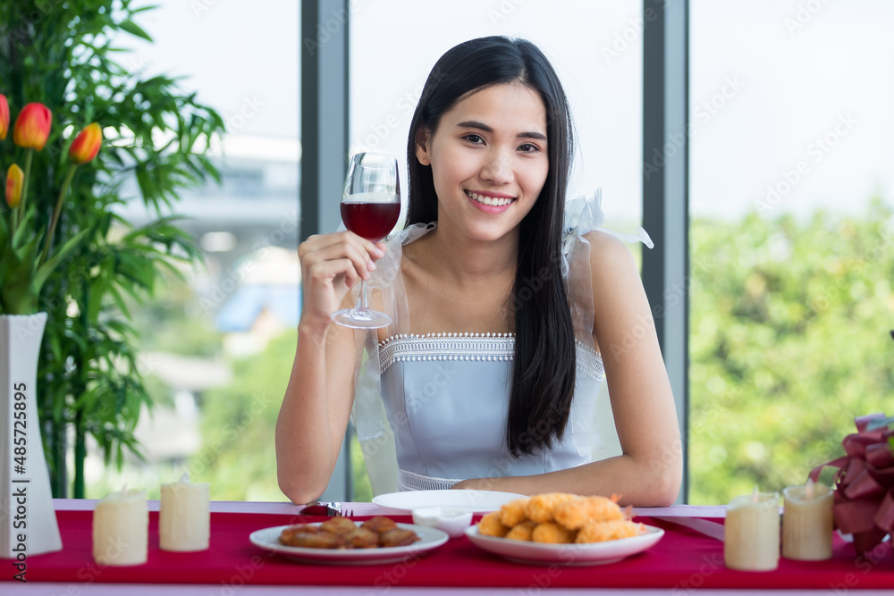 Valentine's day concept, Asian young girl sitting at a table food with wine glasses and bouquet of red and pink roses wine and waiting for her man at in the restaurant background