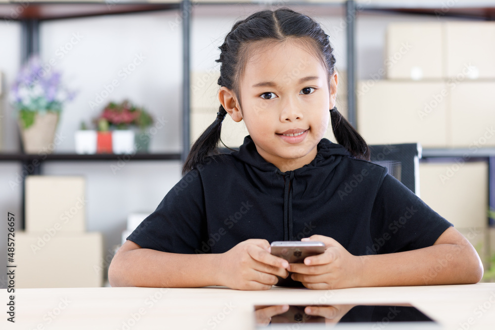 Portrait shot of Asian young cute happy primary schoolgirl in casual outfit sitting at working table smiling holding smartphone in hands. Portrait shot of Asian young cute happy primary schoolgirl in casual outfit sitting at working table smiling holding smartphone in hands.