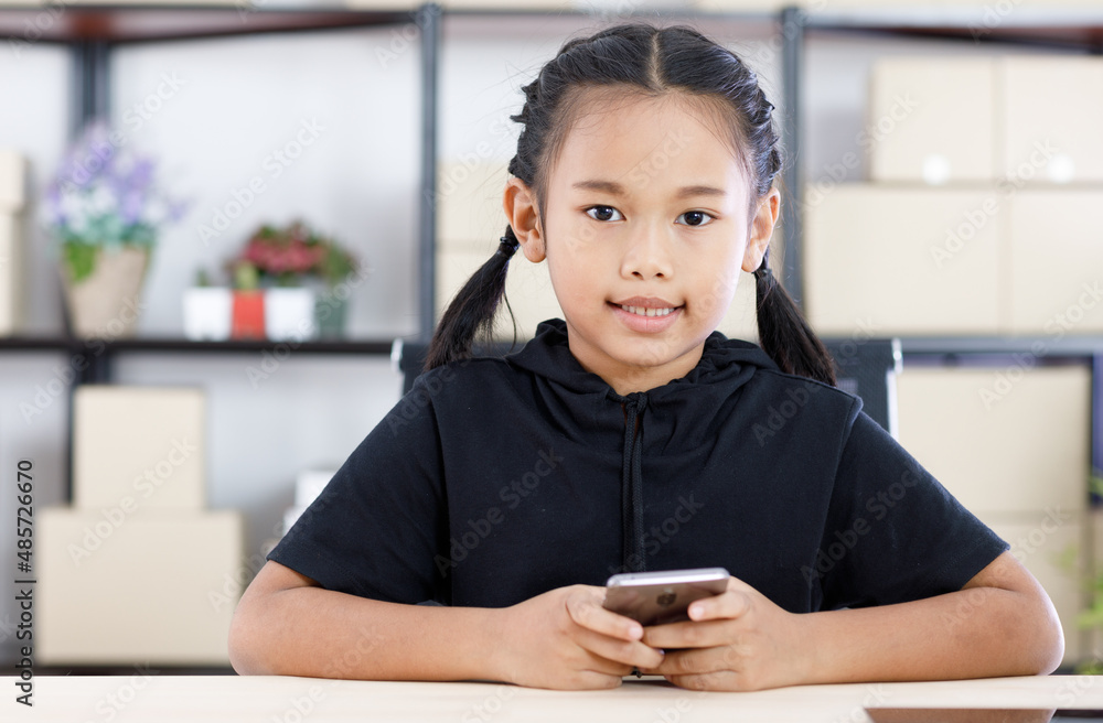 Portrait shot of Asian young cute happy primary schoolgirl in casual outfit sitting at working table smiling holding smartphone in hands. Portrait shot of Asian young cute happy primary schoolgirl in casual outfit sitting at working table smiling holding smartphone in hands.