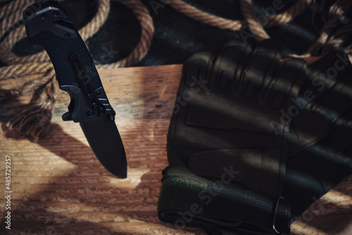 Folding knife, tactical military gloves and rope on a wooden background.
