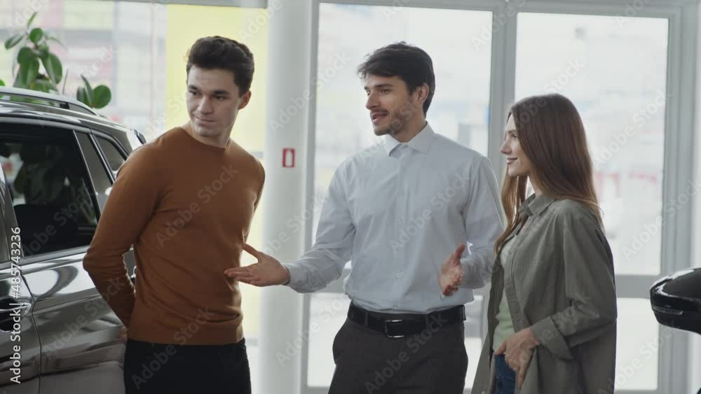Professional sales agent consulting couple of customers at car showroom, explaining benefits of vehicles, tracking shot Professional sales agent consulting couple of customers at car showroom, explaining benefits of vehicles, tracking shot