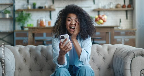 Excited african american woman winner celebrating mobile victory at living room, showing yes gesture. Overjoyed female using smartphone winning gift or prize in social media app, on sofa at home.