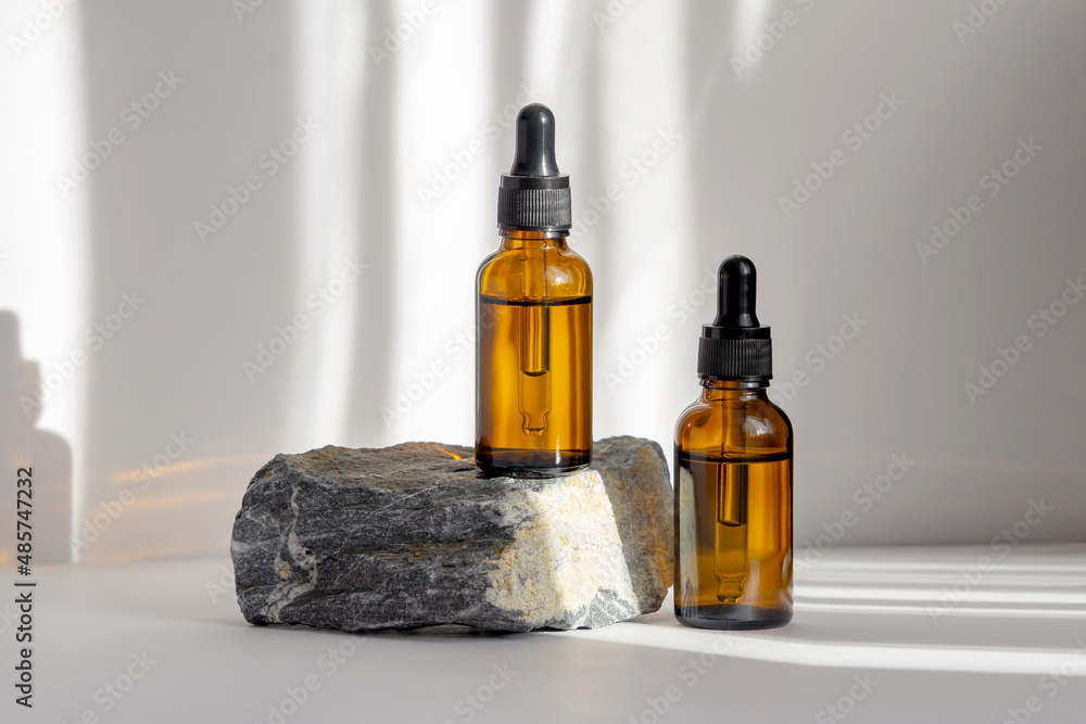 Two mockups of glass bottles with dropper lids on stand made of natural stone. Rays of sunlight fall on objects and on white isolated background. Concept of 3d podium for product demonstration
