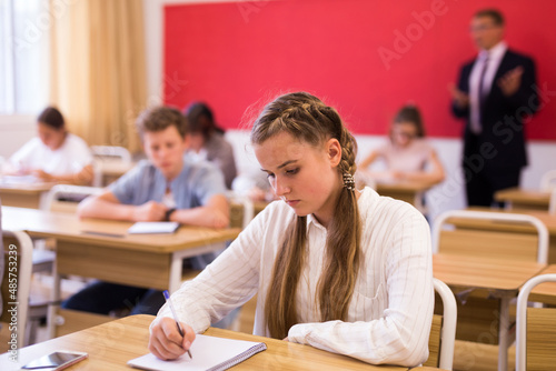 Attractive smart teen girl studying in classroom, listening to lecturer and writing in notebook