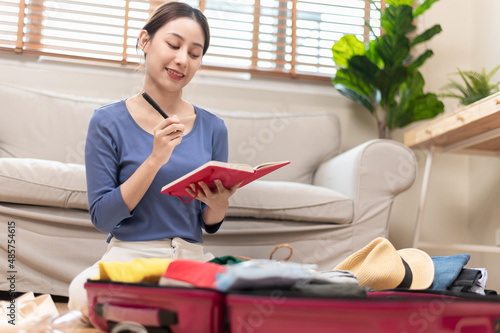 Young beautiful Asian woman using checklist prepare arrange clothes and equipment before go travel oversea after covid pandemic. New normal travel lifestyle concept.