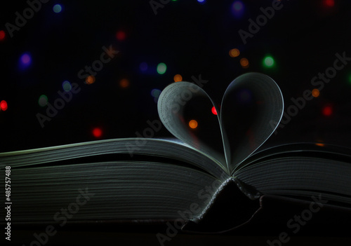 Book pages in shape of heart on dark background with colorful lights. Greering card. Close up, copy space photo