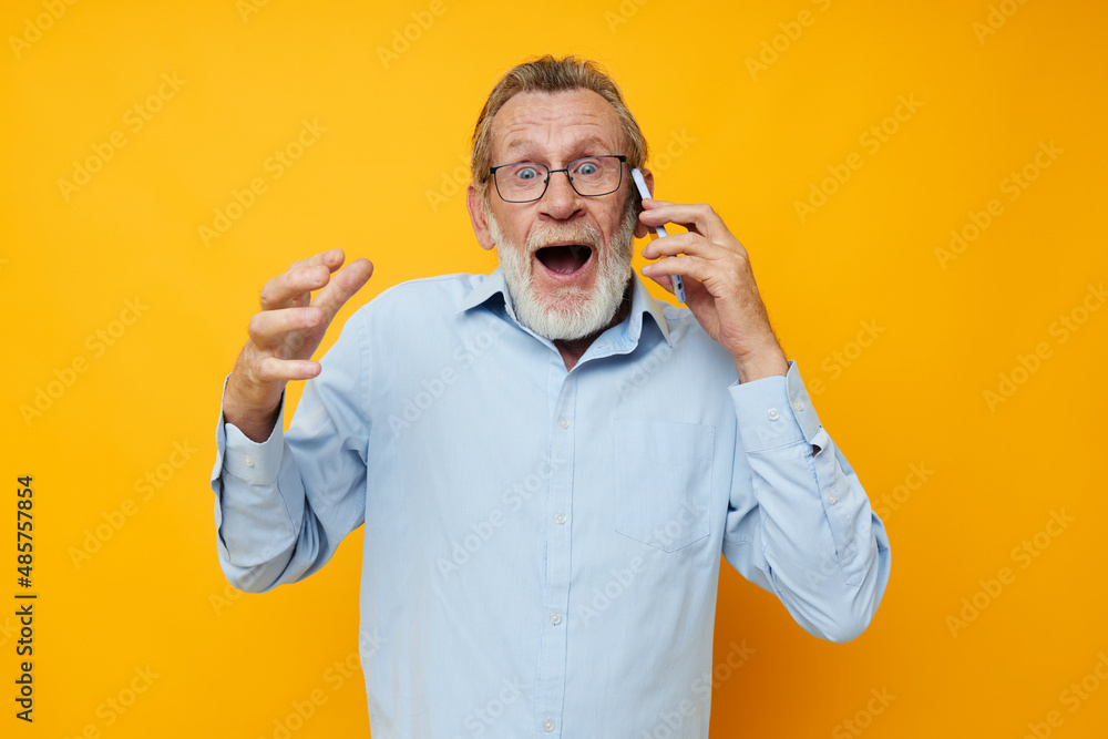 Portrait of happy senior man in a blue shirt and glasses talking on the phone yellow background Portrait of happy senior man in a blue shirt and glasses talking on the phone yellow background